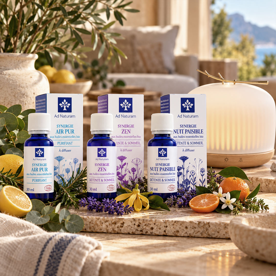 Four Ad Naturam essential oil bottles and boxes arranged on a stone surface outdoors, surrounded by lemon slices, lavender, flowers, greenery, and a diffuser lamp in the background.