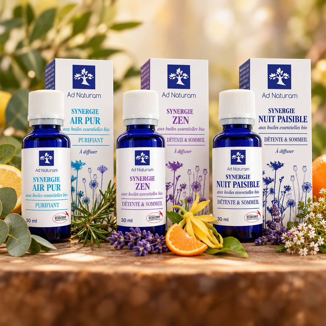 Four Ad Naturam essential oil bottles and their corresponding boxes arranged on a wooden surface with botanical and citrus props. The bottles are cobalt blue with white caps and white labels, and the boxes are white with blue branding and lavender illustrations.