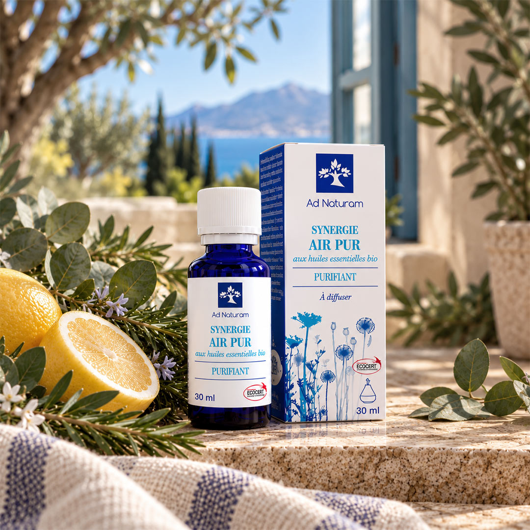 A bottle and box of Ad Naturam Synergie Air Pur diffuser oil placed on a stone surface outdoors, surrounded by greenery, flowers, and lemon slices, with a blurred mountain and blue sky in the background.