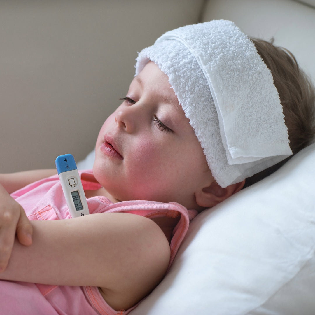 Child lying down with a white towel on head as a compress and digital thermometer in hand