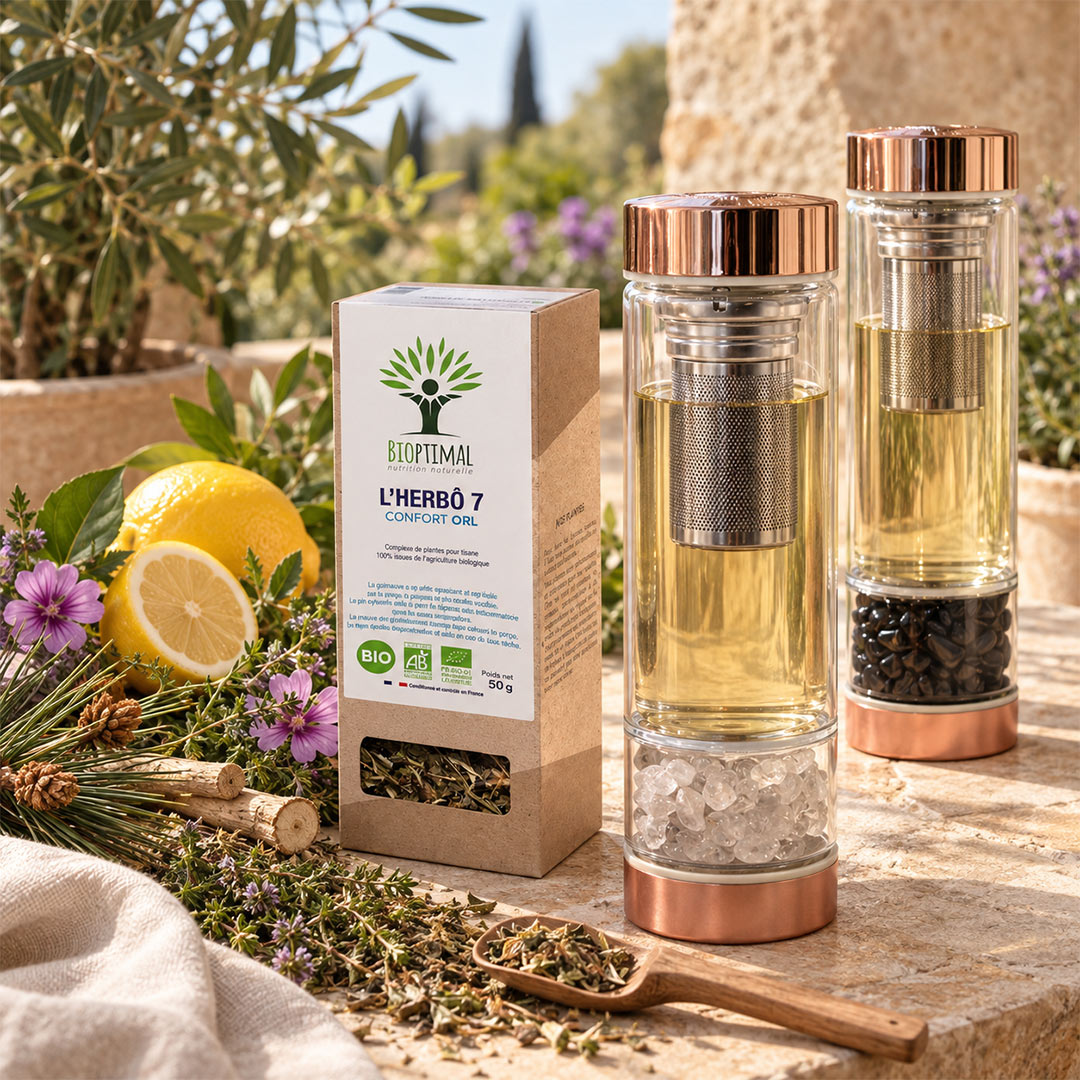 Two clear glass teabottles with copper lids and metal infusers, shown beside a branded product box and a wooden scoop of loose tea on a stone surface, with lemons, herbs, flowers, and potted plants in the background.