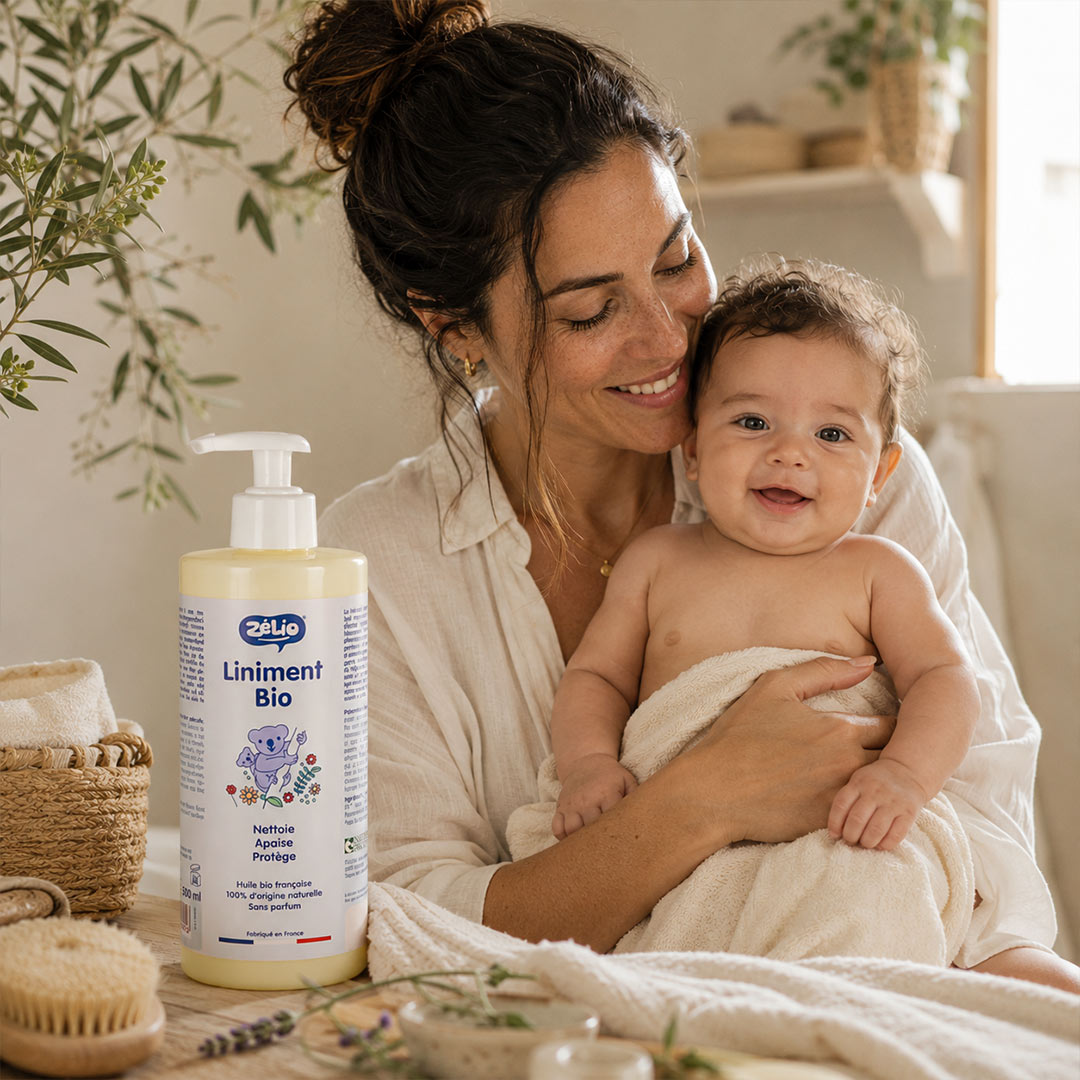 A woman in a white robe holds a baby wrapped in a white towel while promoting a bottle of Liniment Bio. The scene is set in a warm, cozy, rustic bathroom with natural light, wooden shelves, and botanical elements.