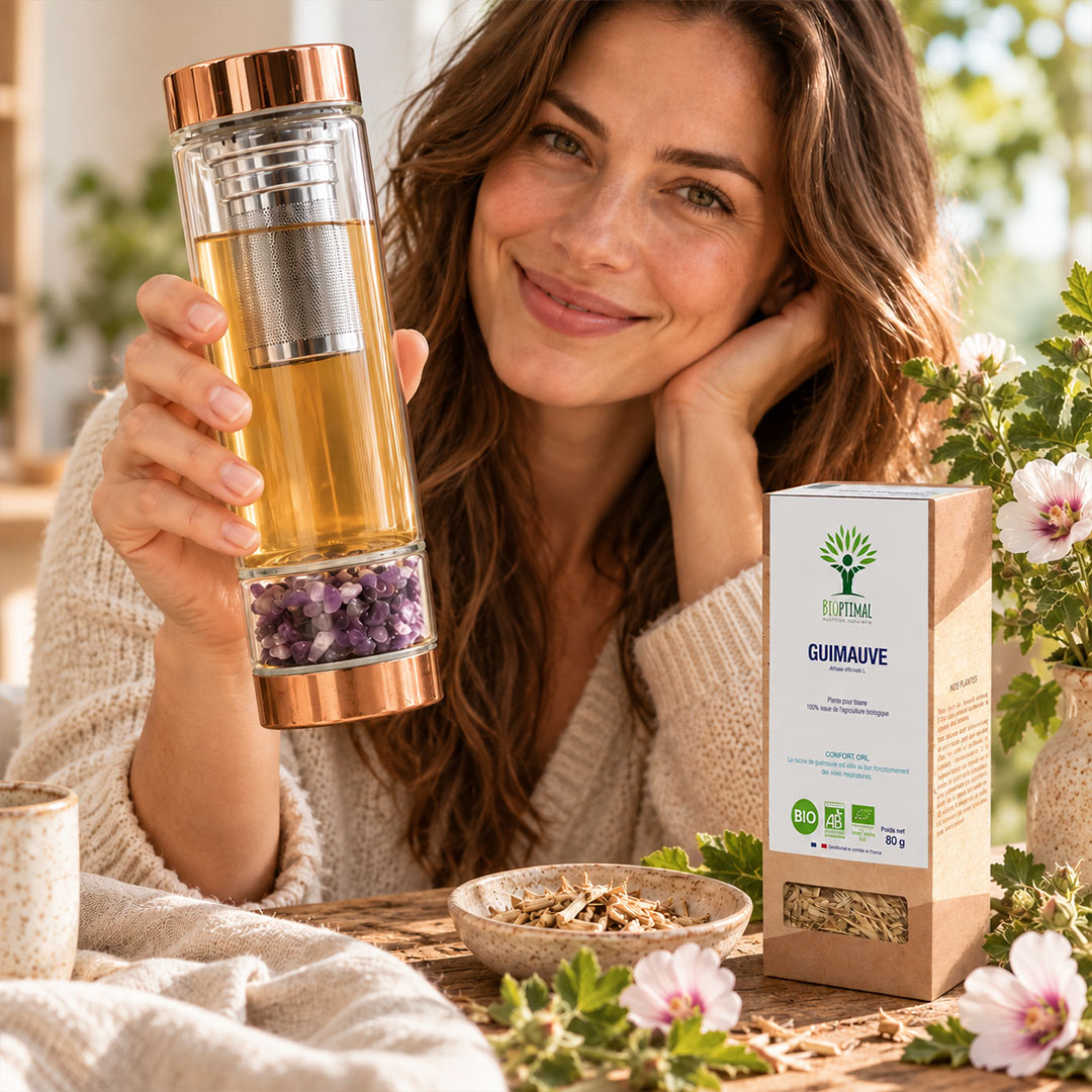 A woman holds a clear glass teapot with copper-toned lid and a visible purple amethyst infuser, standing beside a box labeled 'GUIMAUVE' and a small bowl of loose herbal material on a table. The scene is styled with flowers and natural light in an outdoor indoor setting.