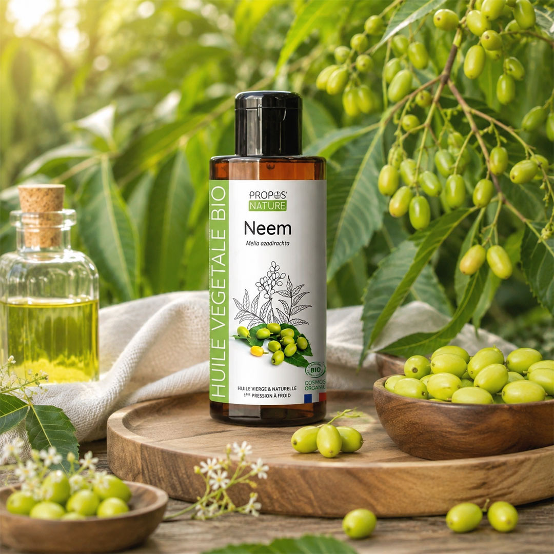 A bottle of Propos Natures Neem vegetable oil stands on a wooden tray surrounded by neem leaves and green neem fruits, with a small glass bottle of oil and bowls of neem fruits in a natural outdoor setting.