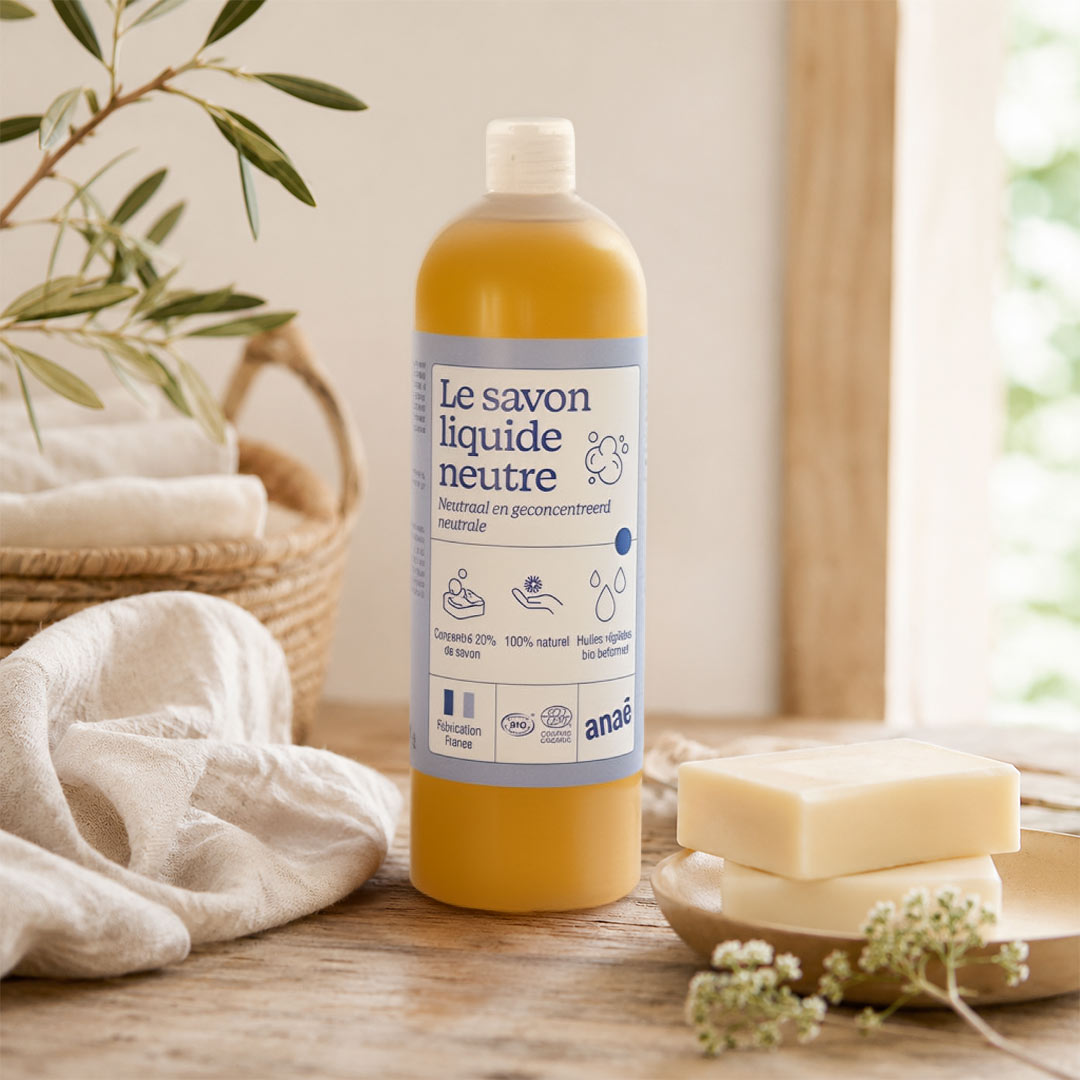 A bottle of Anaâ Le savon liquide neutre (neutral liquid soap) filled with yellow-orange liquid, styled on a wooden table with a bar of soap, a folded cream towel, and green leaves in the background.