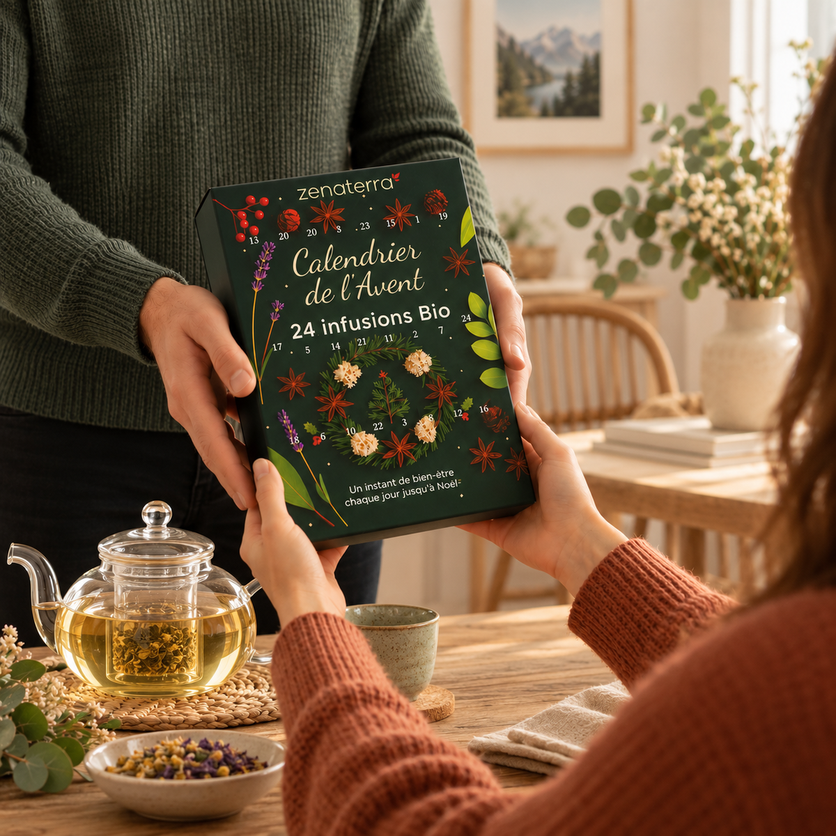 A person hands a green advent calendar titled 'Calendrier de l'Avent 24 Infusions Bio' to another person seated at a wooden table with a glass teapot of herbal tea and dried flowers.