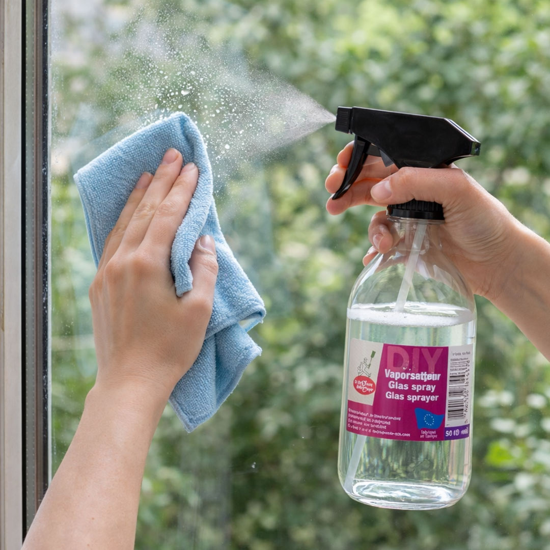 Person cleaning a glass surface with a DIY glass spray and cloth, with greenery in the background.