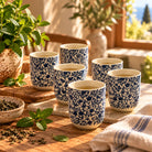 Six ceramic tea cups with a blue and white floral scroll pattern, arranged on wooden boards on a rustic wooden table with mint leaves and a bowl of loose tea.