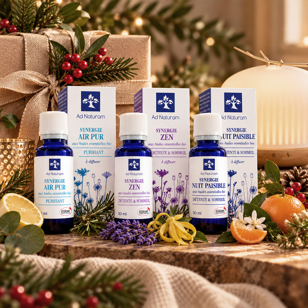 Four Ad Naturum essential oil bottles and their matching cartons arranged on a rustic wooden surface with holiday decorations including pine branches, wrapped gifts, berries, lavender, citrus slices, and a warm blurred background.