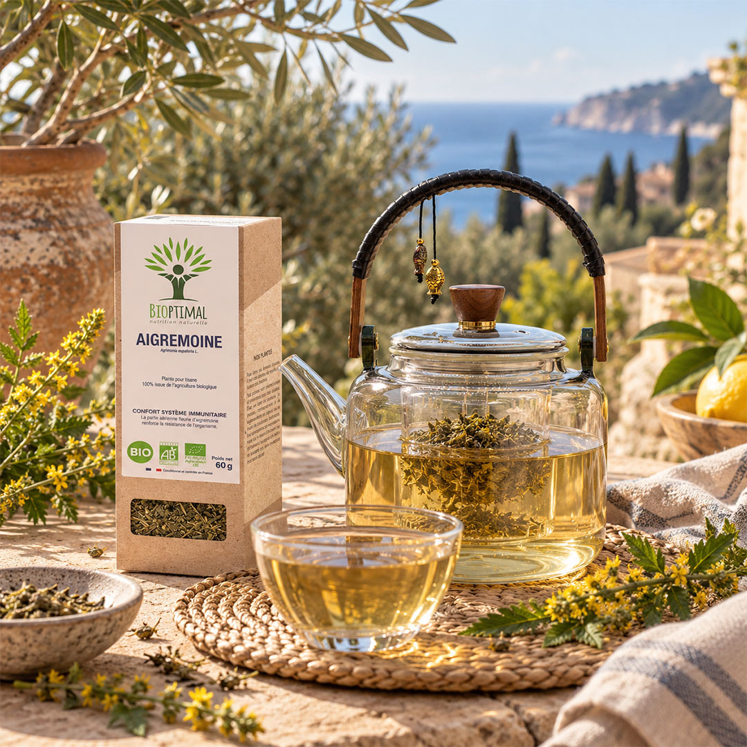 Organic agrimonia tea set displayed on a rustic stone table with a glass teapot, a glass cup of golden tea, and its kraft paper box. The background features a scenic Mediterranean view of the blue sea and cliffs, with olive trees and a vase of flowers.