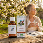 A bottle and box of Herbiolys Gemmotherapy 'Pommier' (apple tree) herbal supplement are displayed on a rustic wooden table outdoors, with a woman holding a cup in the background among blooming apple trees.