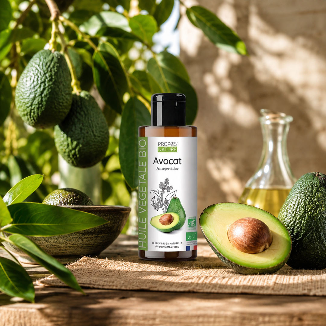 A bottle of organic avocado vegetable oil stands on a woven mat outdoors, surrounded by fresh avocados, green leaves, and a small glass carafe with oil. The label reads 'avocat' and 'Huile Vegetale Bio' with the brand 'agricole bio'.