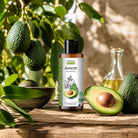 A bottle of organic avocado vegetable oil stands on a woven mat outdoors, surrounded by fresh avocados, green leaves, and a small glass carafe with oil. The label reads 'avocat' and 'Huile Vegetale Bio' with the brand 'agricole bio'.