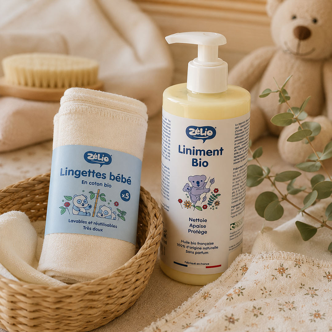 Baby care products arranged on a soft beige fabric surface: a rolled white cotton baby towel with a blue label reading 'Lingettes bébé' stands beside a pump bottle of 'Liniment Bio' with a pale yellow liquid. A woven basket, teddy bear, brush, and greenery are used as styled props.
