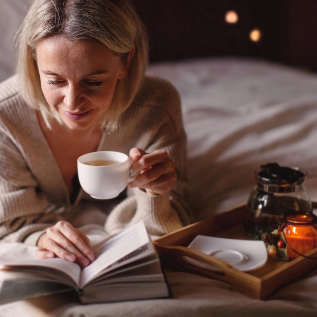 A woman laying on bed with a cup of tea and reading a book