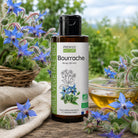 A bottle of Propos Nature Bio Bourrache oil stands on a rustic wooden surface outdoors, surrounded by blooming blue borage flowers, a wicker basket, a glass bowl of golden oil, and a soft blurred green garden background.