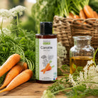 A bottle of PROPOS'S NATURE Carotte (carrot) organic macerate oil stands on a rustic wooden table surrounded by fresh carrots, green herbs, and white carrot flowers, with a glass bottle of the oil nearby.