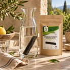 A kraft paper pouch labeled “BÂTON DE CHARBON VEGETAL ACTIF” stands beside a clear glass carafe filled with water on a sunlit stone surface, with lemon, plants, and a rustic outdoor setting in the background.