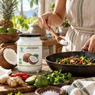 A jar of organic coconut oil sits on a rustic kitchen table next to a person wearing a striped apron, who is holding a wooden spoon filled with salt over a pan of cooked vegetables. Fresh limes, ginger, and chili peppers are scattered on the table.
