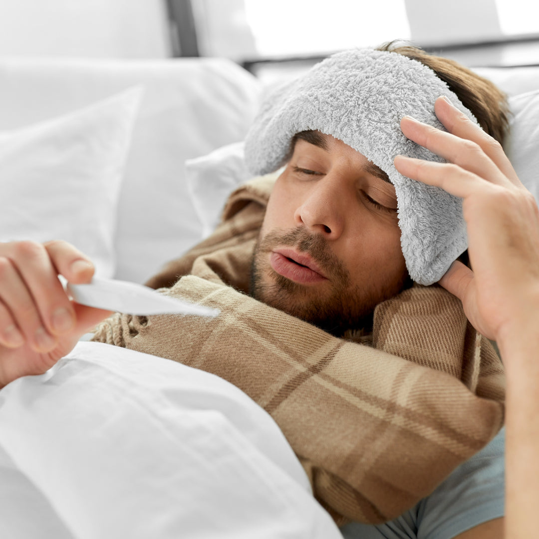 A man laying in bed with a cold having a cloth on the forehead and looking at the termometer