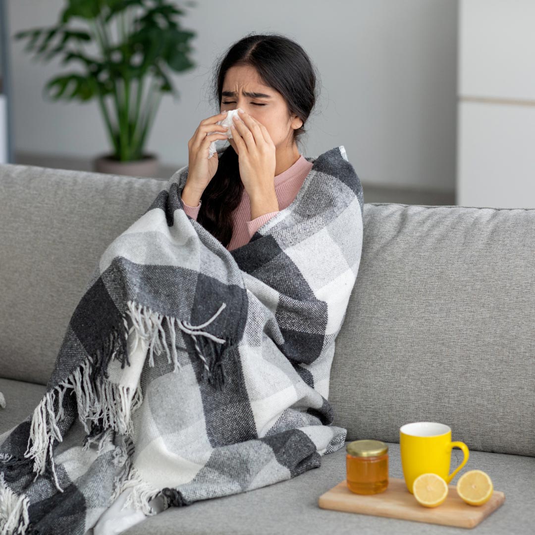 A woman sitting in a grey sofa with a blanket around herself, blowing her nose, having a cold