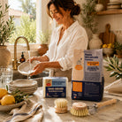 Woman rinsing a ceramic plate in a bright Mediterranean kitchen with the Natural Dishwashing Starter Kit, including ecological dish soap, dishwasher powder, a wooden dish brush and replacement brush head.