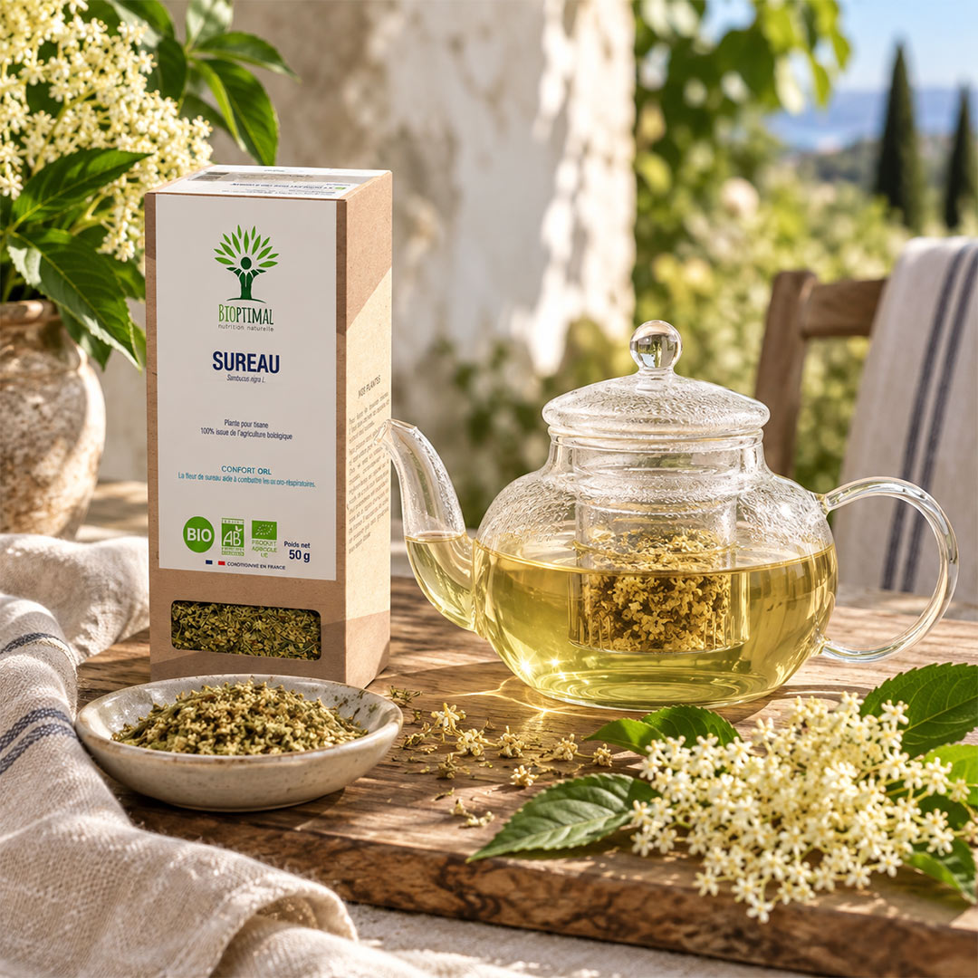 A glass teapot filled with brewed herbal tea sits on a wooden table beside a box of Bioptimal Sureau elderflower tea and a small bowl of loose tea leaves, with elderflower blossoms and greenery in the background.
