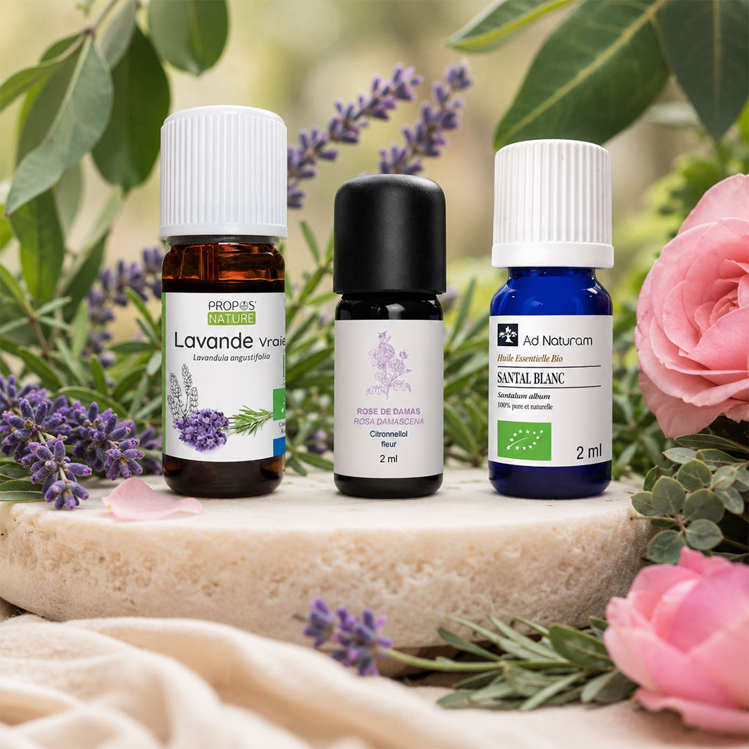 Three essential oil bottles displayed on a stone slab with lavender sprigs and pink roses. The bottles include Propos'Nature Organic Lavander, Essenciagua  Rose Damask, Ad Naturum Sandalwood