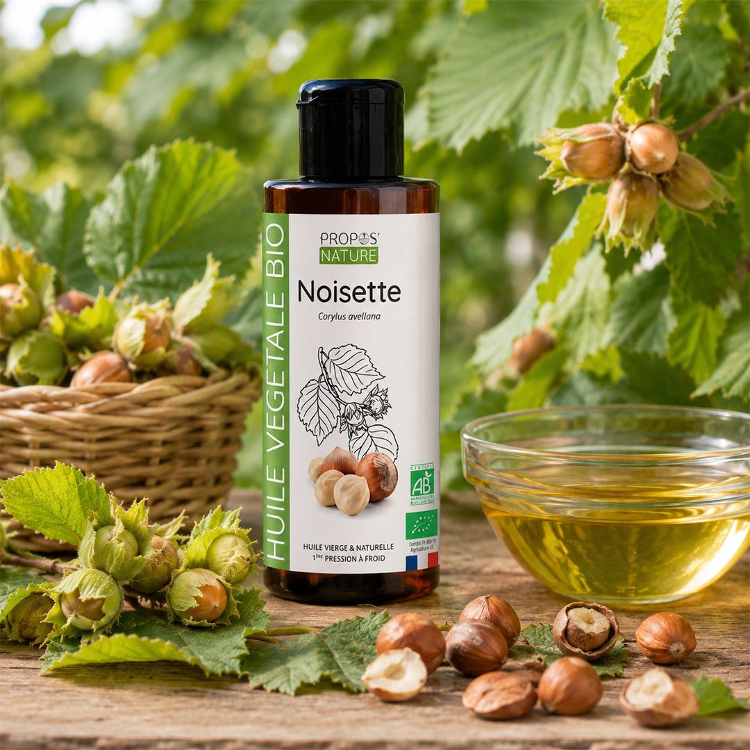 A bottle of Propos Naturel Bio Noisette (hazelnut) vegetable oil is displayed on a rustic wooden table surrounded by fresh hazelnuts, green leaves, and a glass bowl of the golden-yellow oil, set against a lush background of green foliage.