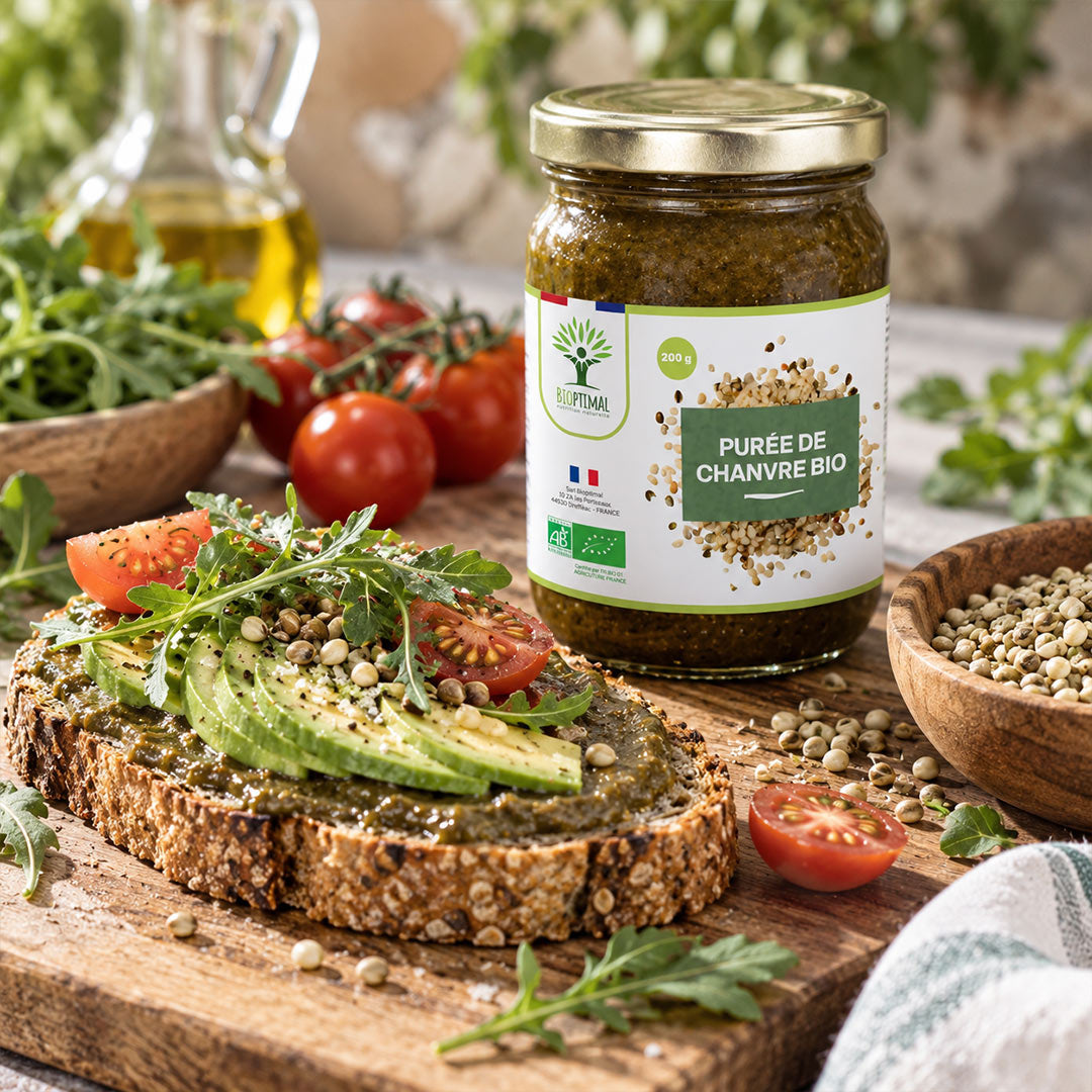 A jar of Bio Purée de Chamvre from the brand Bioptimal is displayed on a wooden board next to an open slice of toasted multigrain bread topped with avocado, tomatoes, and herbs. The background is a rustic kitchen setting with fresh tomatoes, a bottle of olive oil, and bowls of ingredients.