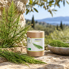 A cylindrical kraft paper jar labeled 'PRÈLE BIO' with a white botanical label sits outdoors on a stone surface beside fresh green herb sprigs and a bowl, with a blurred olive garden landscape in the background.