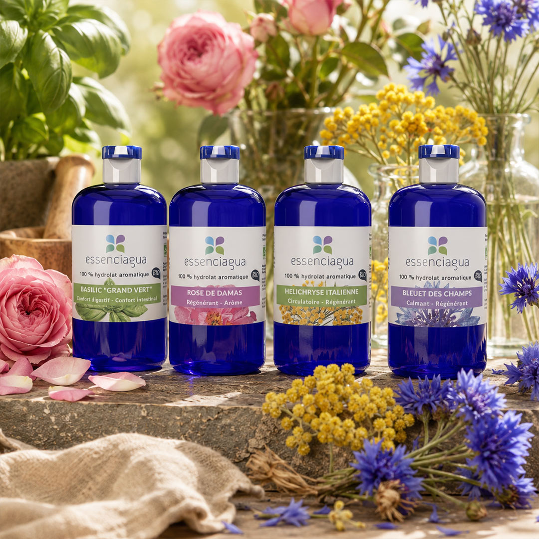 Four Essenciaguahydrosol bottles in cobalt blue glass with white labels, arranged on a rustic stone surface with pink and yellow flowers in the background.