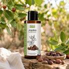 A bottle of Propos Nature Jojoba vegetable oil stands on a rustic wooden surface outdoors, surrounded by green leaves, small red berries, and a small bowl of jojoba nuts. The bottle has a black cap and a white label with black text and botanical illustrations.