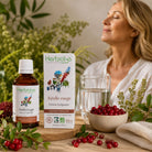 A woman in a white robe relaxes beside a bottle and box of Herbialys Herbophorée Airelle rouge tincture, with cranberries, a glass of water, and greenery on a wooden table.