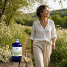 A woman in a white linen shirt and beige pants walks along a sunlit path surrounded by wildflowers, with a blue bottle of Essenciagua Meadowsweet hydrosol placed on a wooden table in the foreground.