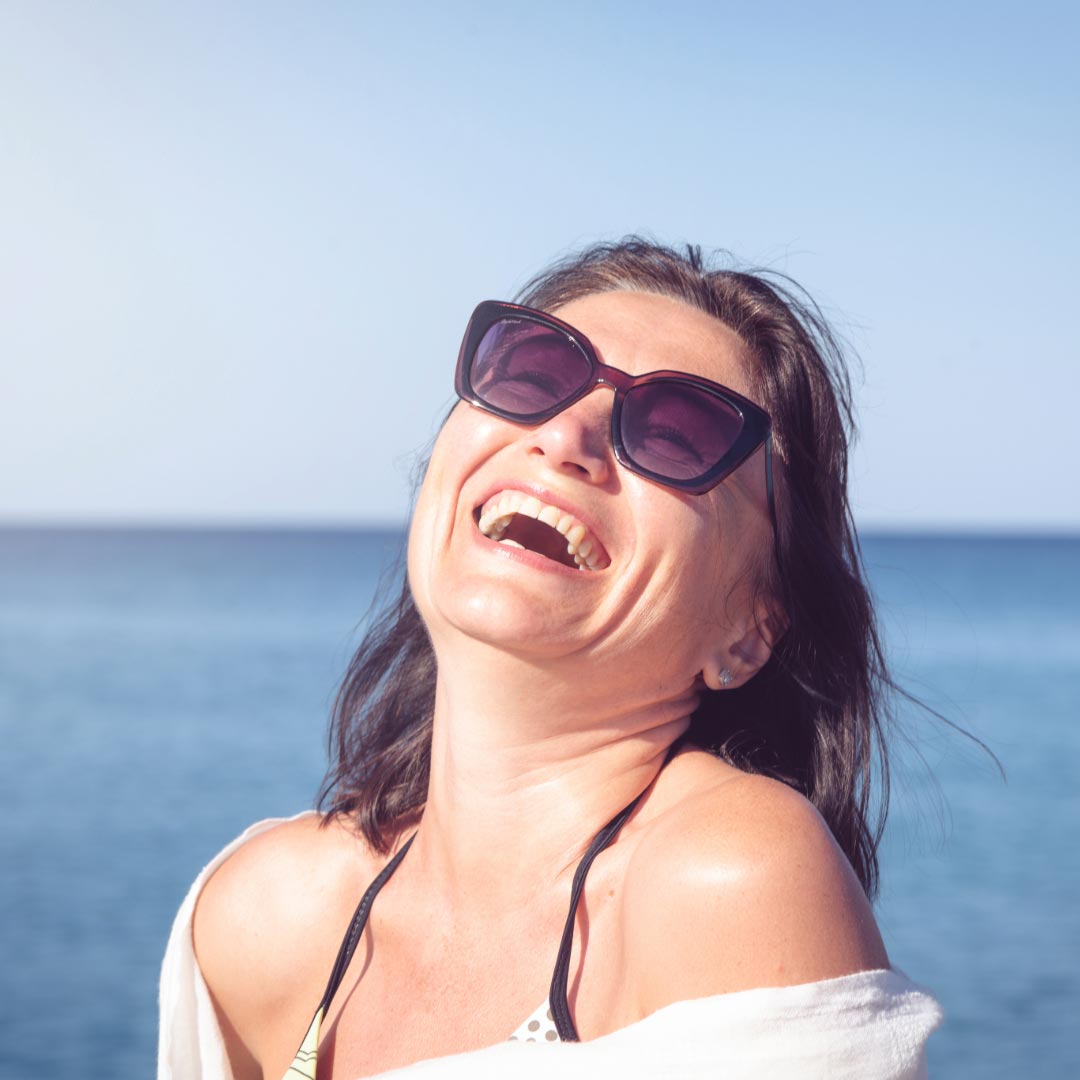 A woman laughing at the beach wearing sunglasses and bikini top with a shirt over
