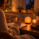 A warm, cozy indoor scene with a person in a knitted sweater writing at a wooden table. On the table are a glowing Himalayan Moon salt lamp, a small candle holder, a mug, and a notebook.