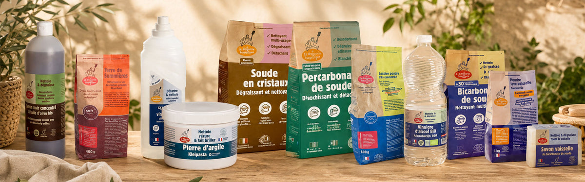 A collection of natural soap and cleaning products arranged on a wooden table with a rustic background of green leaves and warm sunlight. The products include various bars, bottles, and boxes from brands like Pierre d'ague and Percarbonbe de soude.