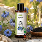 A bottle of Propos Nature Nigelle vegetable oil stands on a rustic wooden surface surrounded by blue flowers Nigella sativa, black seeds in a wooden bowl, and soft neutral fabric. The product label is white with green and orange accents, and the bottle itself is amber brown with a black cap.