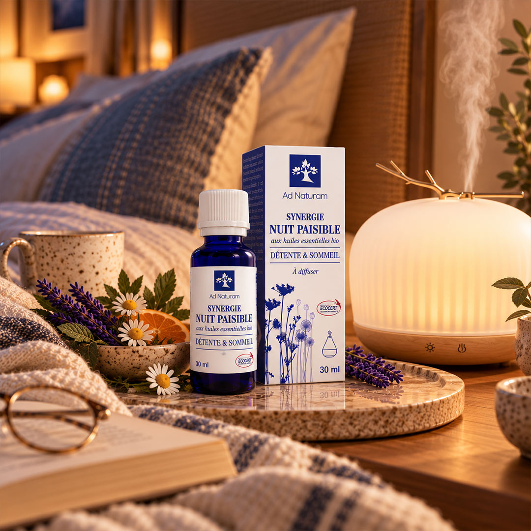 A bottle and box of Ad Naturum Synergie Nuit Paisible diffuser oil are styled on a wooden tray beside a glowing diffuser, lavender, daisies, a mug, and an open book in a cozy bedroom setting.