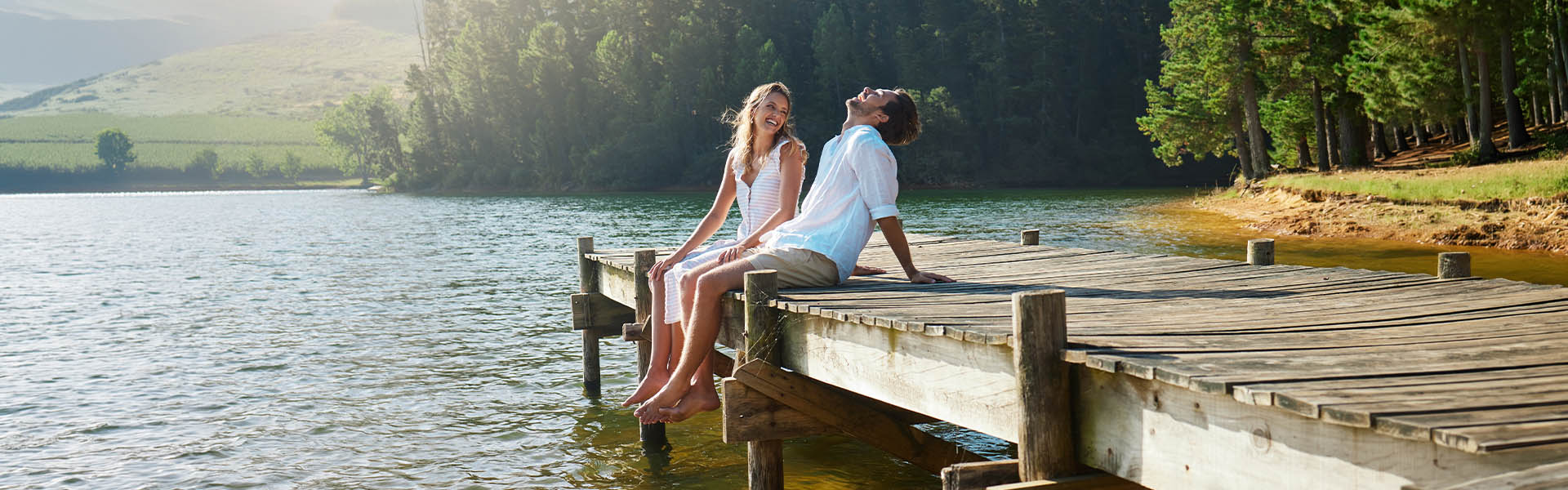 Two people sitting on a wooden dock by a lake with trees in the background