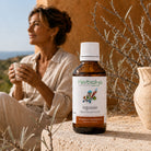 A bottle of Herbiolys Sea Buckthorn gemmotherapy sits on a stone ledge in a warm, rustic setting with a woman holding a mug in the soft-focus background.extract