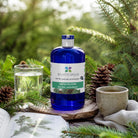 A cobalt blue bottle of Essenciagua Sapin Pictine hydrosol is styled on a wooden surface with silver fir branches, a glass of water, a ceramic mug, and an open book in a natural outdoor setting.