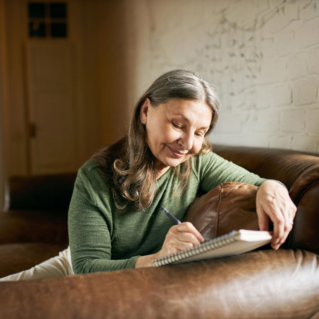 A middle aged woman sitting in a sofa writing in her journal
