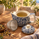 A blue and white floral porcelain teapot with a matching lid and metal strainer, filled with tea and set on a wooden table outdoors with dried flowers and greenery in the background.
