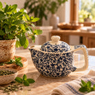 A ceramic teapot with a blue floral pattern on a cream background, styled on a wooden table with mint leaves, a small bowl of dried herbs, and a potted plant in a warm kitchen setting.