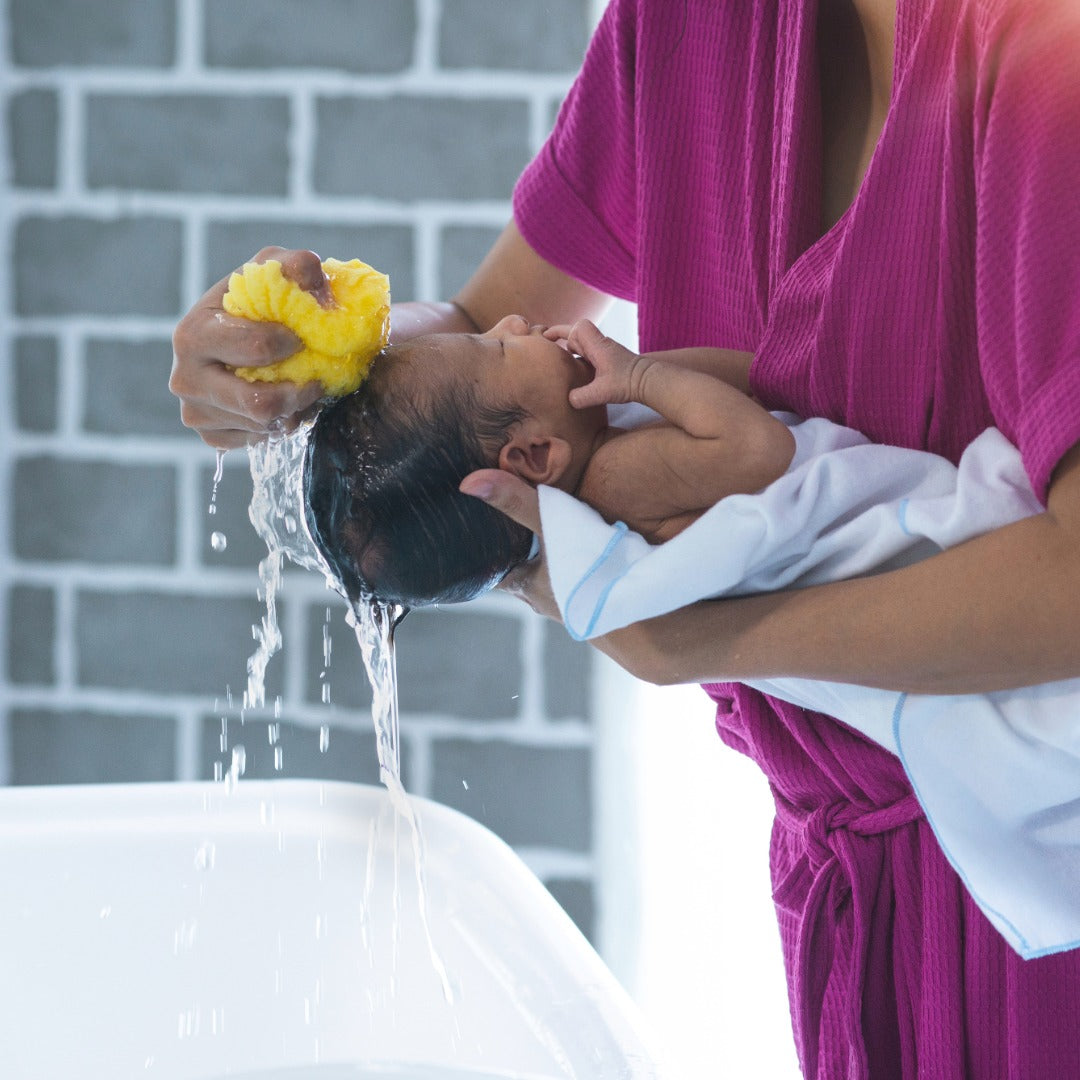 Person washing a baby with water from a yellow sponge against a gray brick wall.