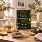 A zenaterra Advent calendar of 24 organic infusions stands on a wooden table surrounded by a glass teapot with golden tea, cups, bowls of loose tea and lavender, and seasonal greenery in a warm, softly lit interior.