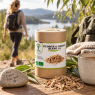 A cylindrical kraft paper container of organic white birch bark labeled 'ÉCORCE DE SAULE BLANC BIO' sits on a wooden table outdoors, with loose birch bark scattered around it. A hiker with a backpack walks blurred in the background near a lake, and a mug, leaves, and a stone are also visible in the scene.