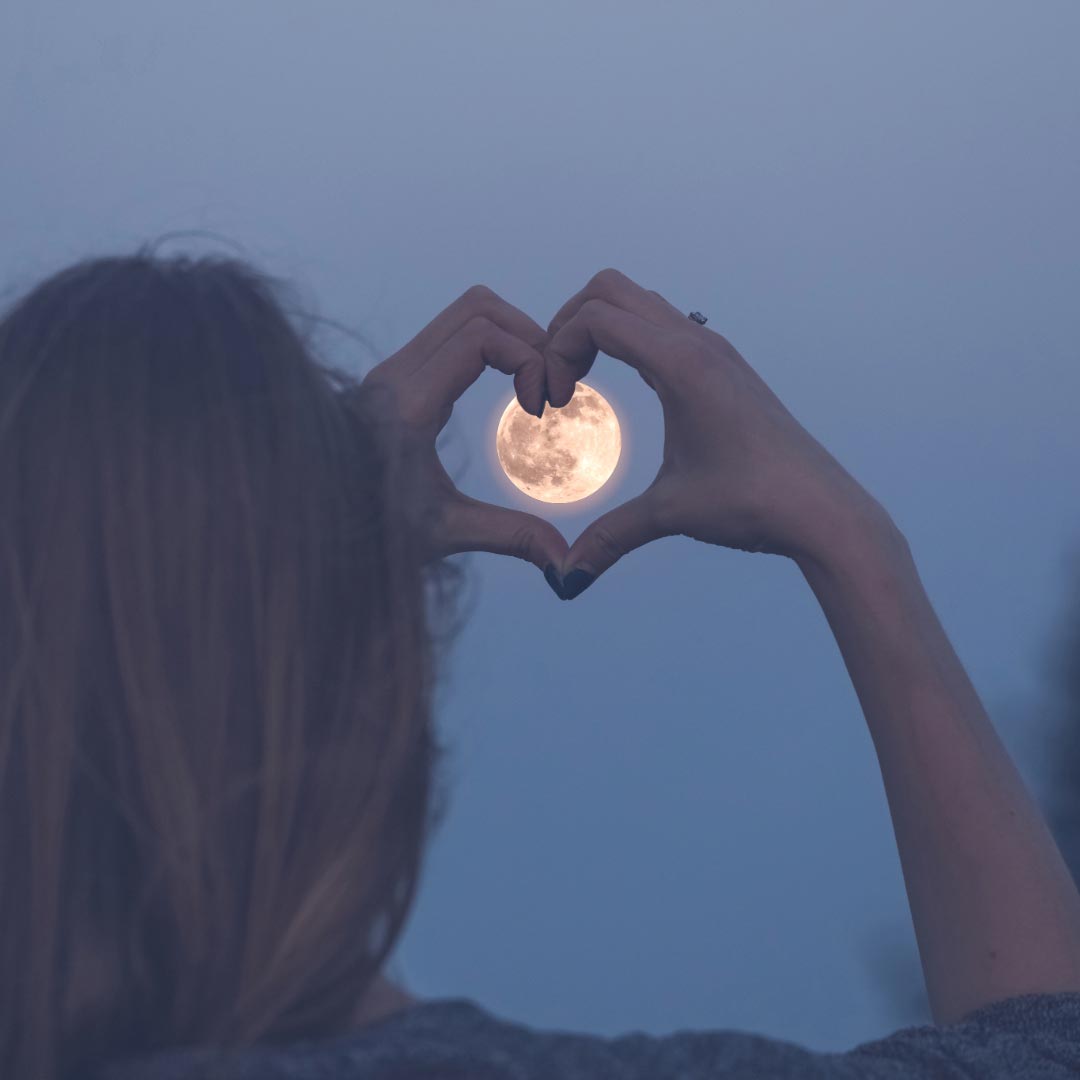 A woman holding her hands in the shape of a heart around the moon